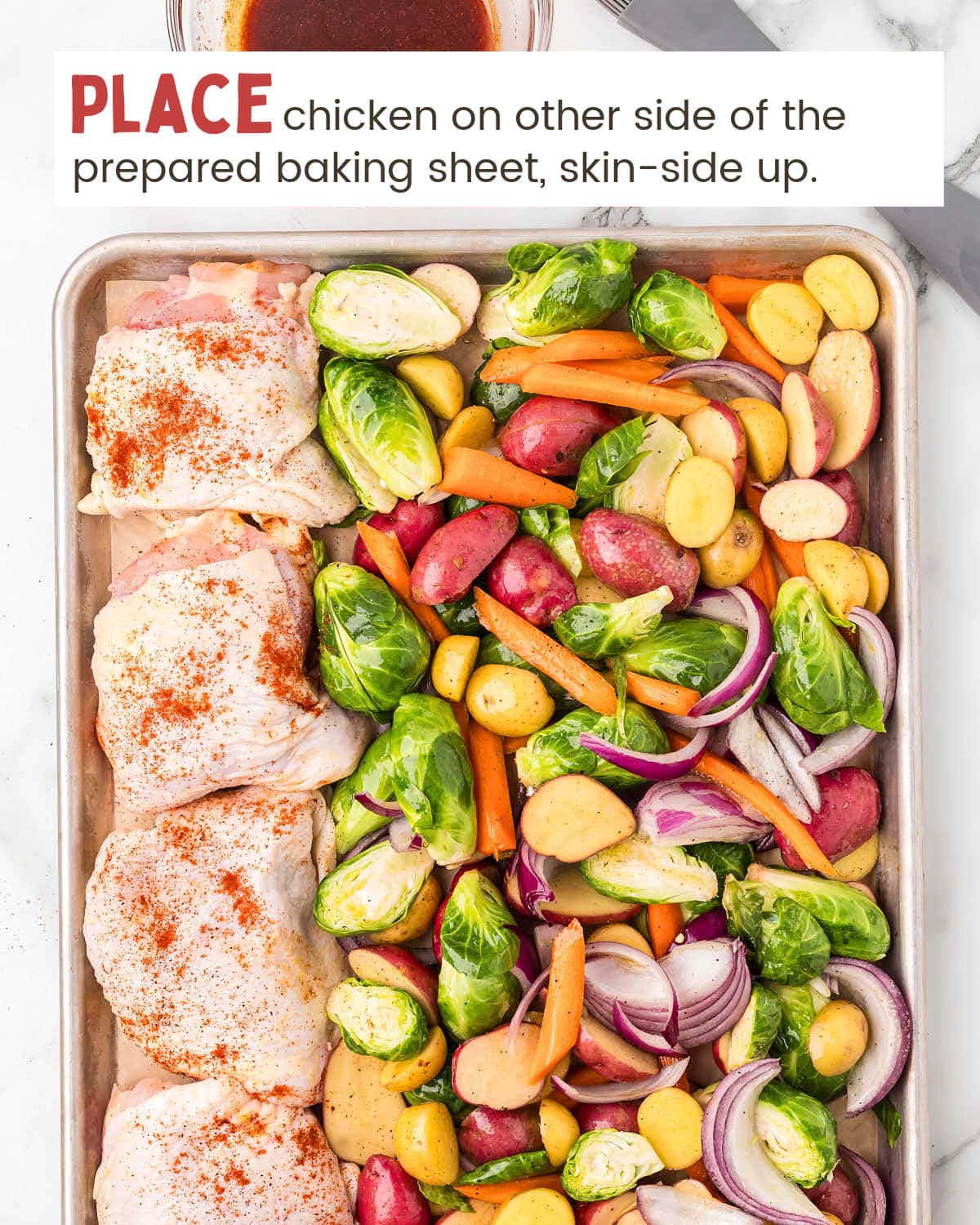 Red onions, baby potatoes, sliced carrots, and Brussels sprouts next to raw chicken thighs on a baking sheet.