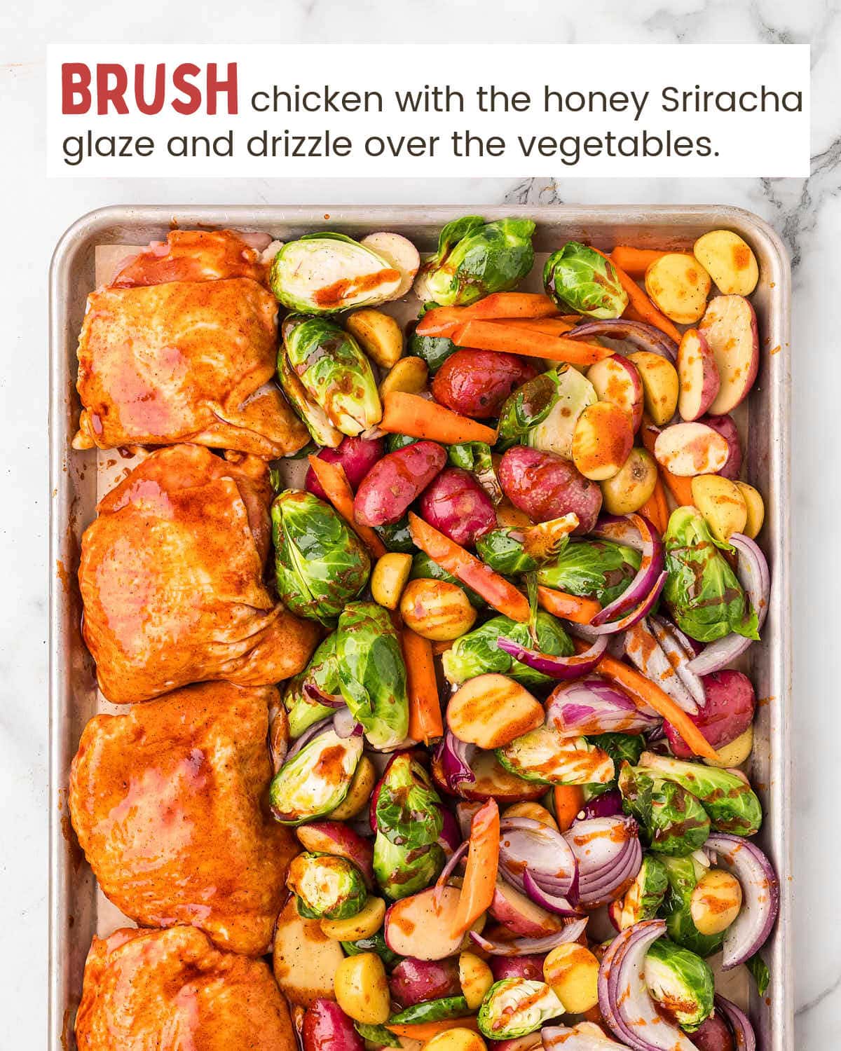 Red onions, baby potatoes, sliced carrots, and Brussels sprouts next to honey siracha chicken thighs on a baking sheet.