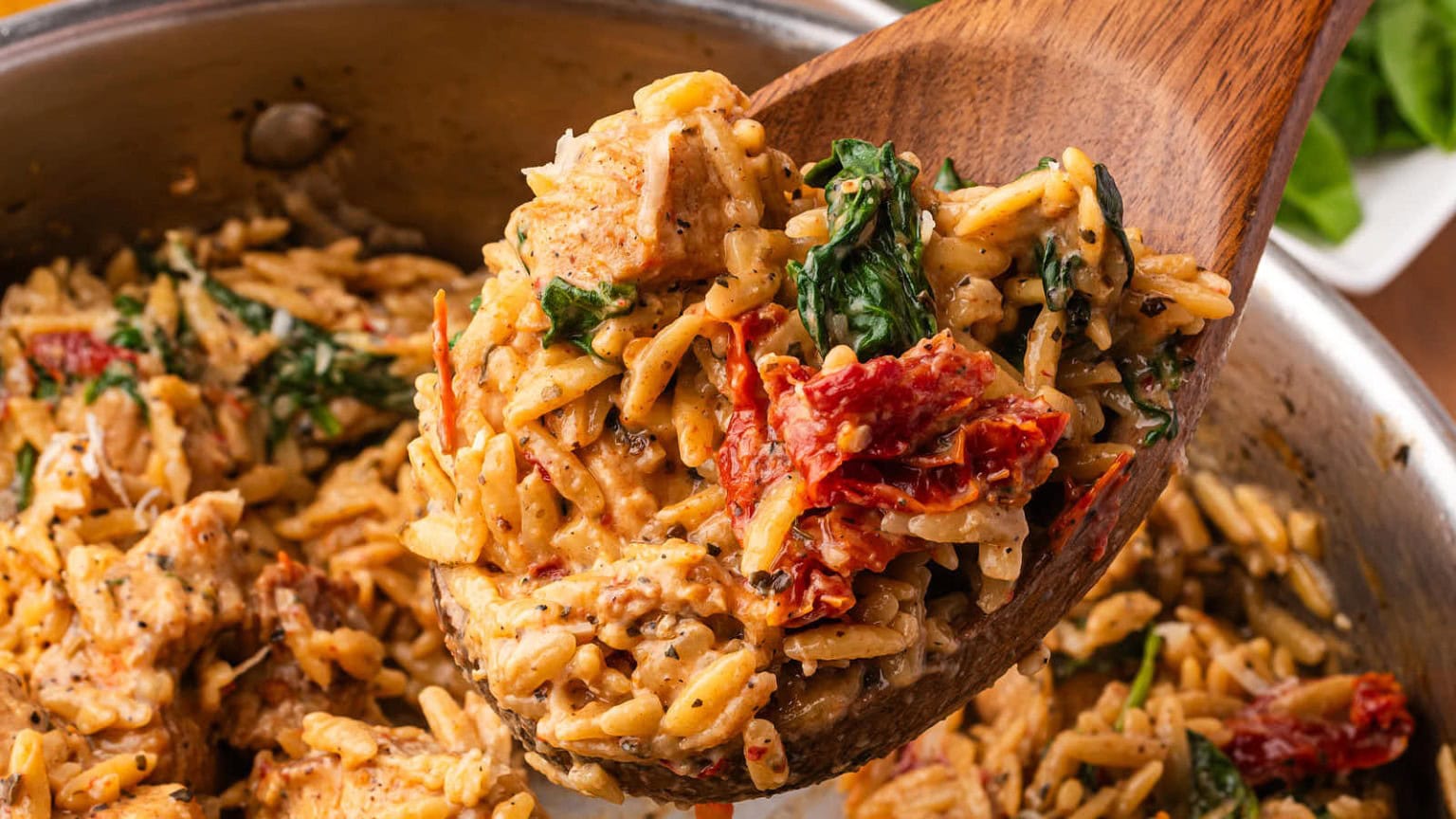 A wooden spoon lifts creamy orzo pasta mixed with chicken, spinach, and sun-dried tomatoes from a stainless steel pot.