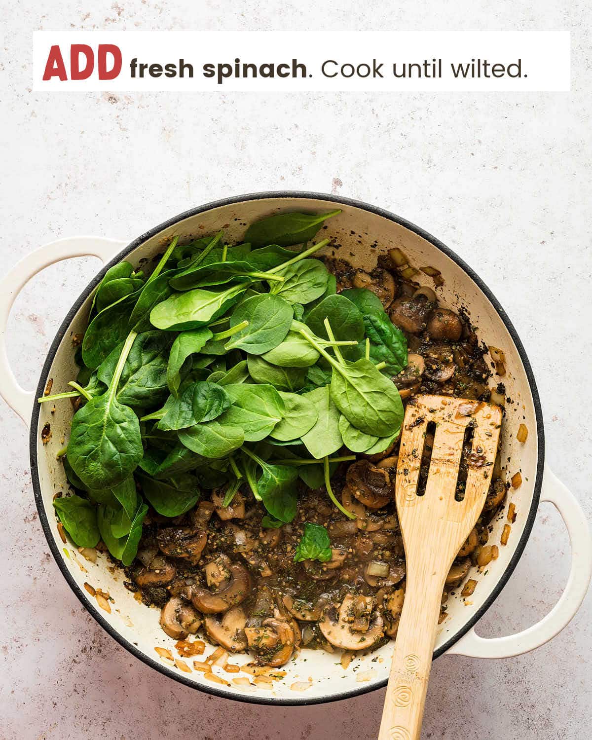Mushrooms, onions, seasonings, and herbs topped with fresh spinach in a pot. A wooden spatula rests in the pot.