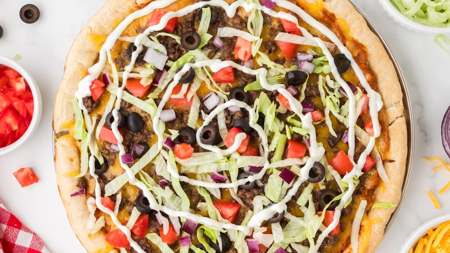A colorful taco pizza topped with lettuce, tomatoes, olives, onions, ground beef, and drizzled with sour cream, served with diced tomatoes.
