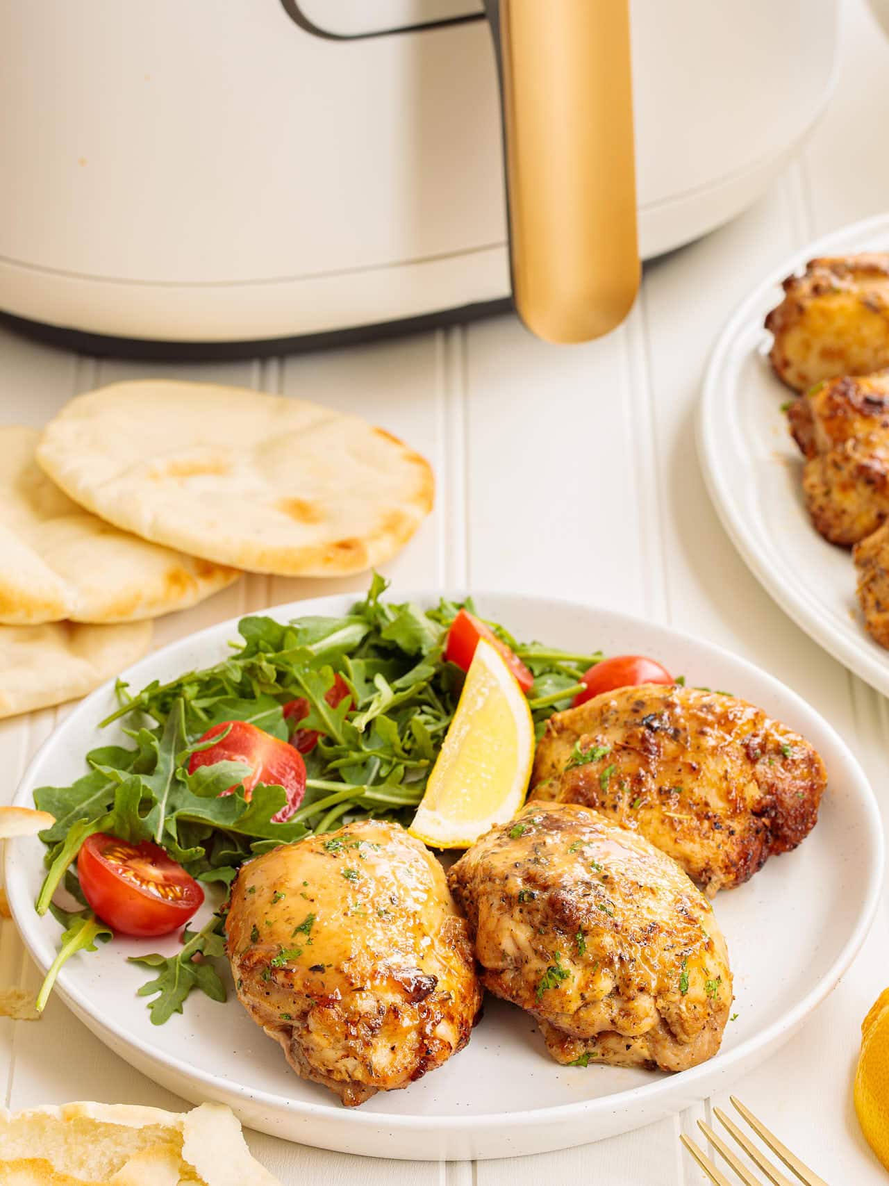 Plate of three air fryer chicken thighs served with arugula, cherry tomatoes, and a lemon wedge, with pita bread and a white air fryer in the background.