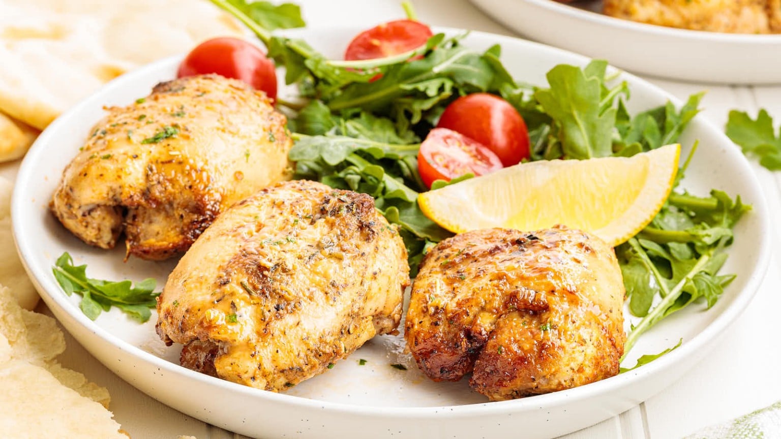 Golden-brown chicken thighs served with cherry tomatoes, arugula, and lemon on a white plate, alongside soft flatbread.