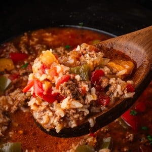 A wooden spoon lifts a hearty scoop of Stuffed Pepper Soup with ground beef, rice, and colorful bell peppers from a slow cooker.