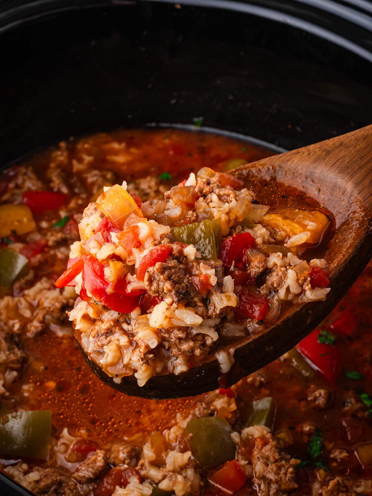 A wooden spoon lifts a hearty scoop of Stuffed Pepper Soup with ground beef, rice, and colorful bell peppers from a slow cooker.