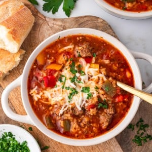 A bowl of Stuffed Pepper Soup topped with melted cheese and chopped parsley, surrounded by fresh bread and herbs on a wooden board.