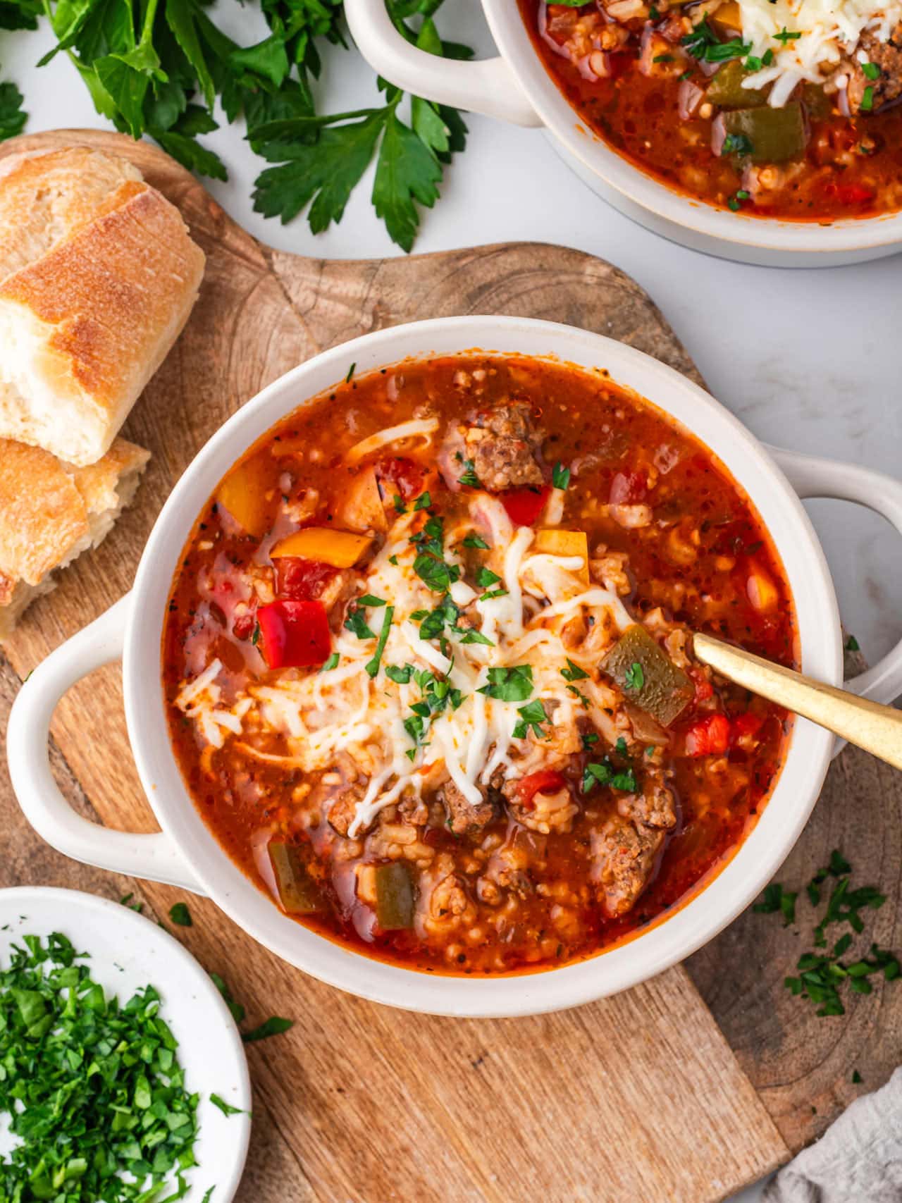A bowl of Stuffed Pepper Soup topped with melted cheese and chopped parsley, surrounded by fresh bread and herbs on a wooden board.