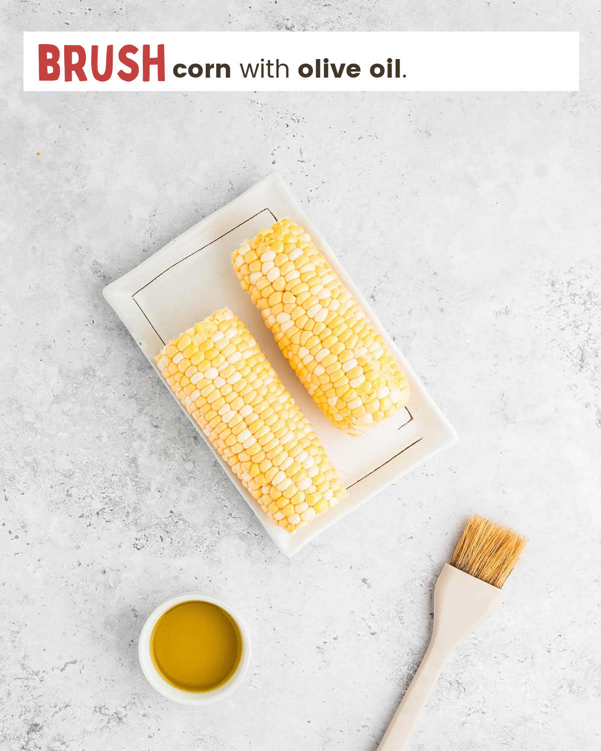 Two ears of corn on a plate, a dish of olive oil, and a brush on a table.