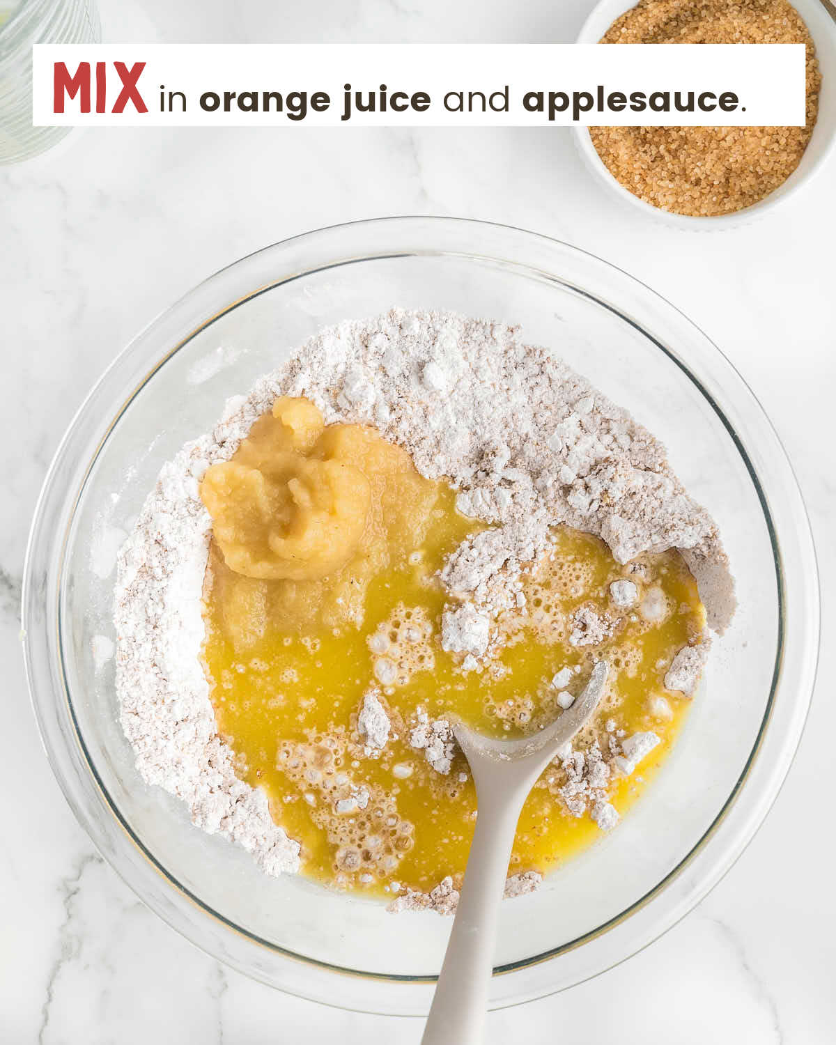 A wooden spoon mixing orange juice and applesauce into a flour mixture in a glass bowl.