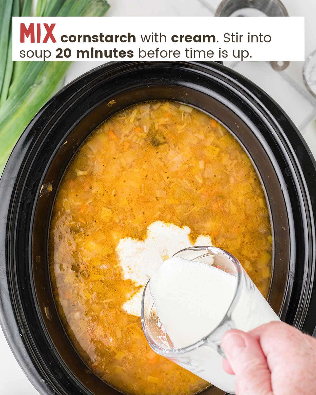 A hand pouring a cornstarch and cream mixture in a measuring cup into A wooden spoon in potato and leek soup in a slow cooker.