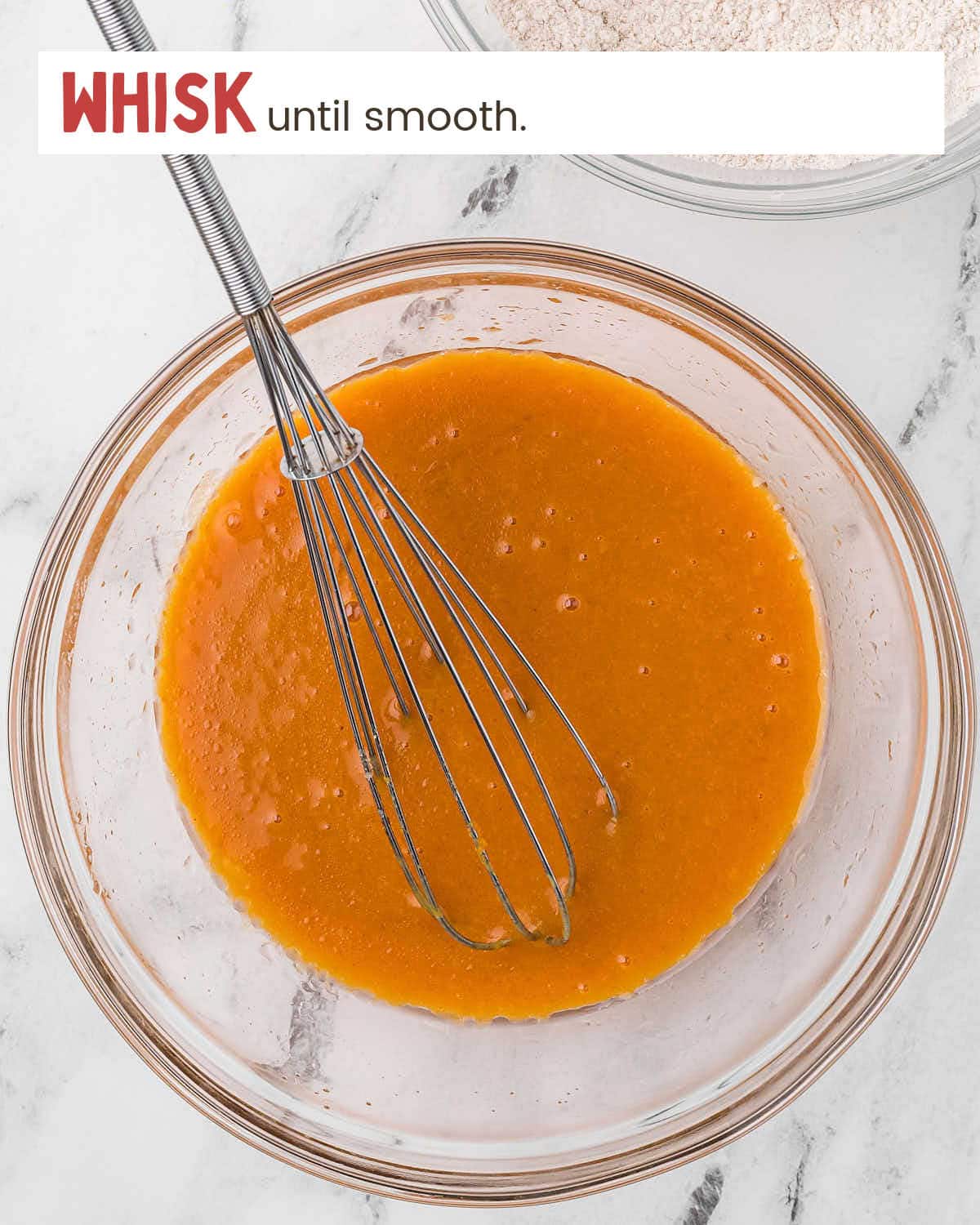 A whisk in whisked pumpkin muffin ingredients in a glass bowl.