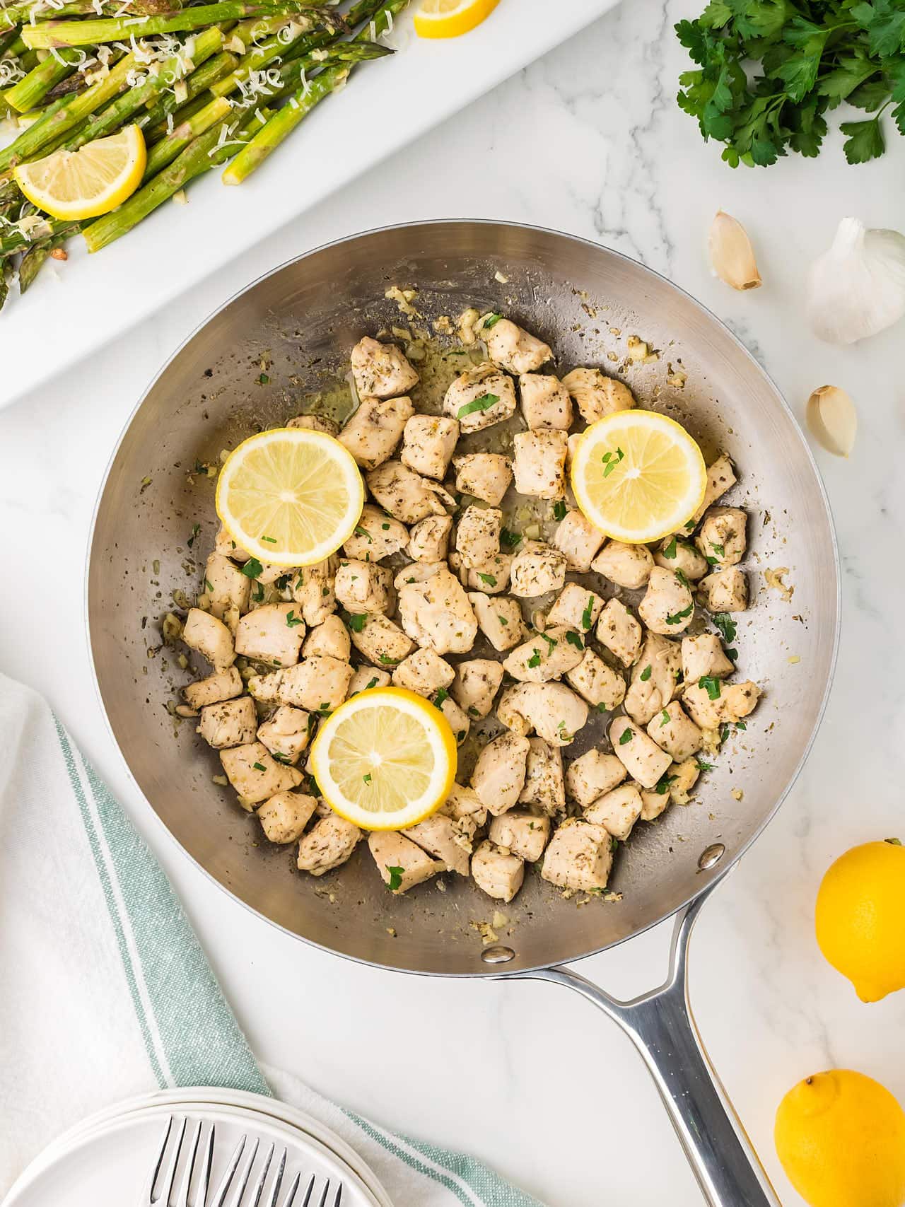 Overhead view of a skillet of Garlic Butter Chicken Bites with lemon slices, next to roasted asparagus.