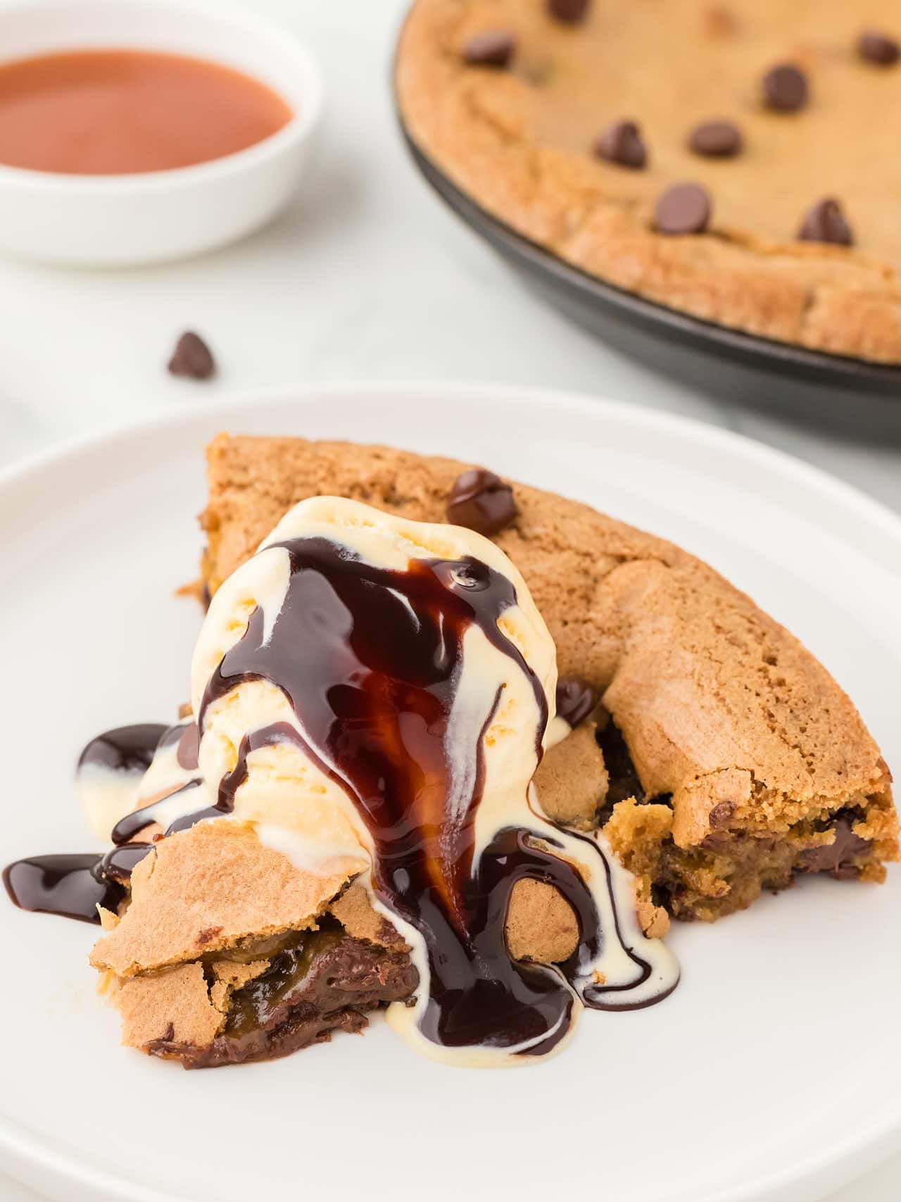 Slice of chocolate chip pizookie topped with vanilla ice cream and rich hot fudge on a white plate.