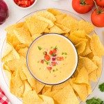 Queso dip topped with tomato, red onion, and cilantro, served with tortilla chips on a platter.