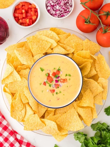 Queso dip topped with tomato, red onion, and cilantro, served with tortilla chips on a platter.