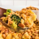A wooden spoon lifting a cheesy serving of Broccoli Cauliflower Casserole topped with golden stuffing crumbs.