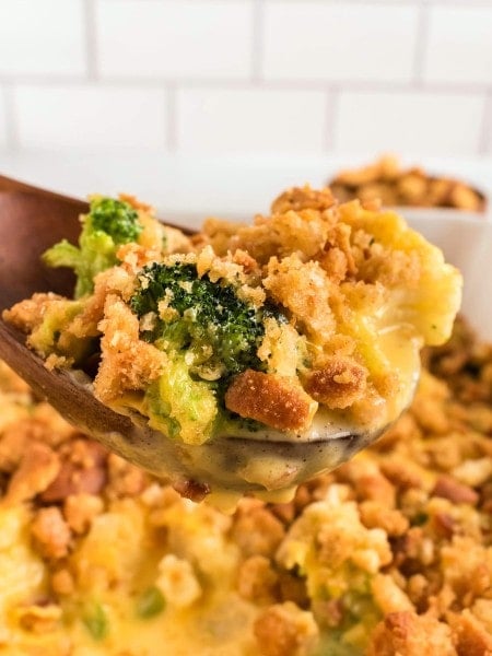 A wooden spoon lifting a cheesy serving of Broccoli Cauliflower Casserole topped with golden stuffing crumbs.