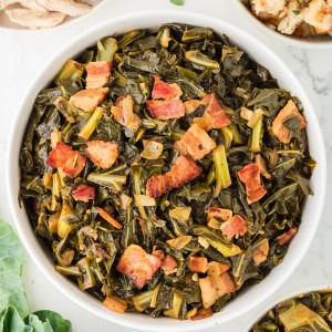 Collard greens cooked with crispy bacon, onions, and garlic served in a white bowl on a marble table, surrounded by stuffing, turkey, and fresh greens.