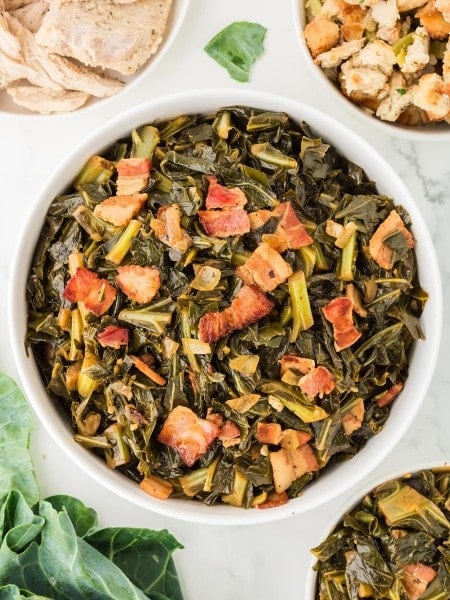 Collard greens cooked with crispy bacon, onions, and garlic served in a white bowl on a marble table, surrounded by stuffing, turkey, and fresh greens.