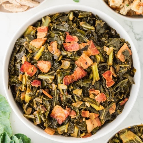 Collard greens cooked with crispy bacon, onions, and garlic served in a white bowl on a marble table, surrounded by stuffing, turkey, and fresh greens.