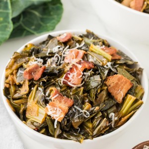 Collard Greens topped with crispy bacon and shredded cheese in a white bowl, with fresh greens in the background.