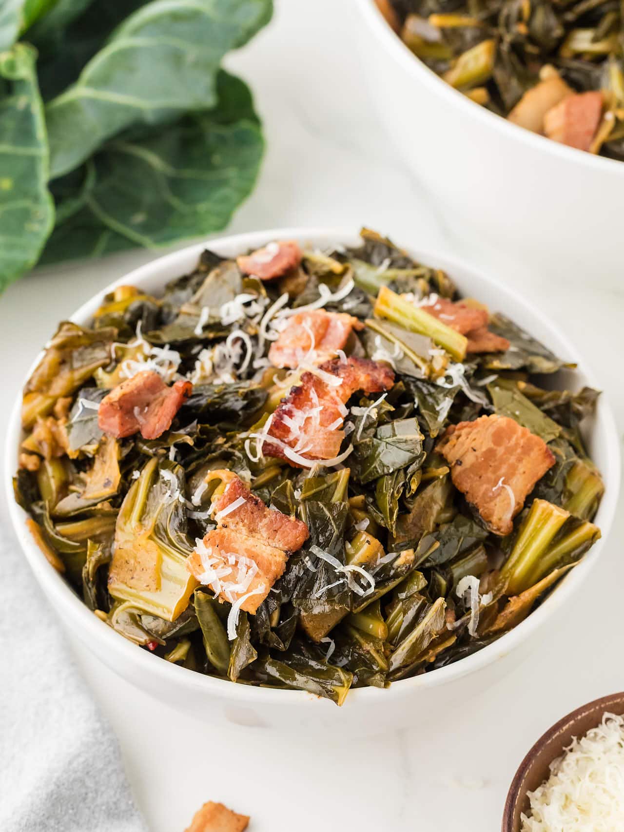 Collard Greens topped with crispy bacon and shredded cheese in a white bowl, with fresh greens in the background.