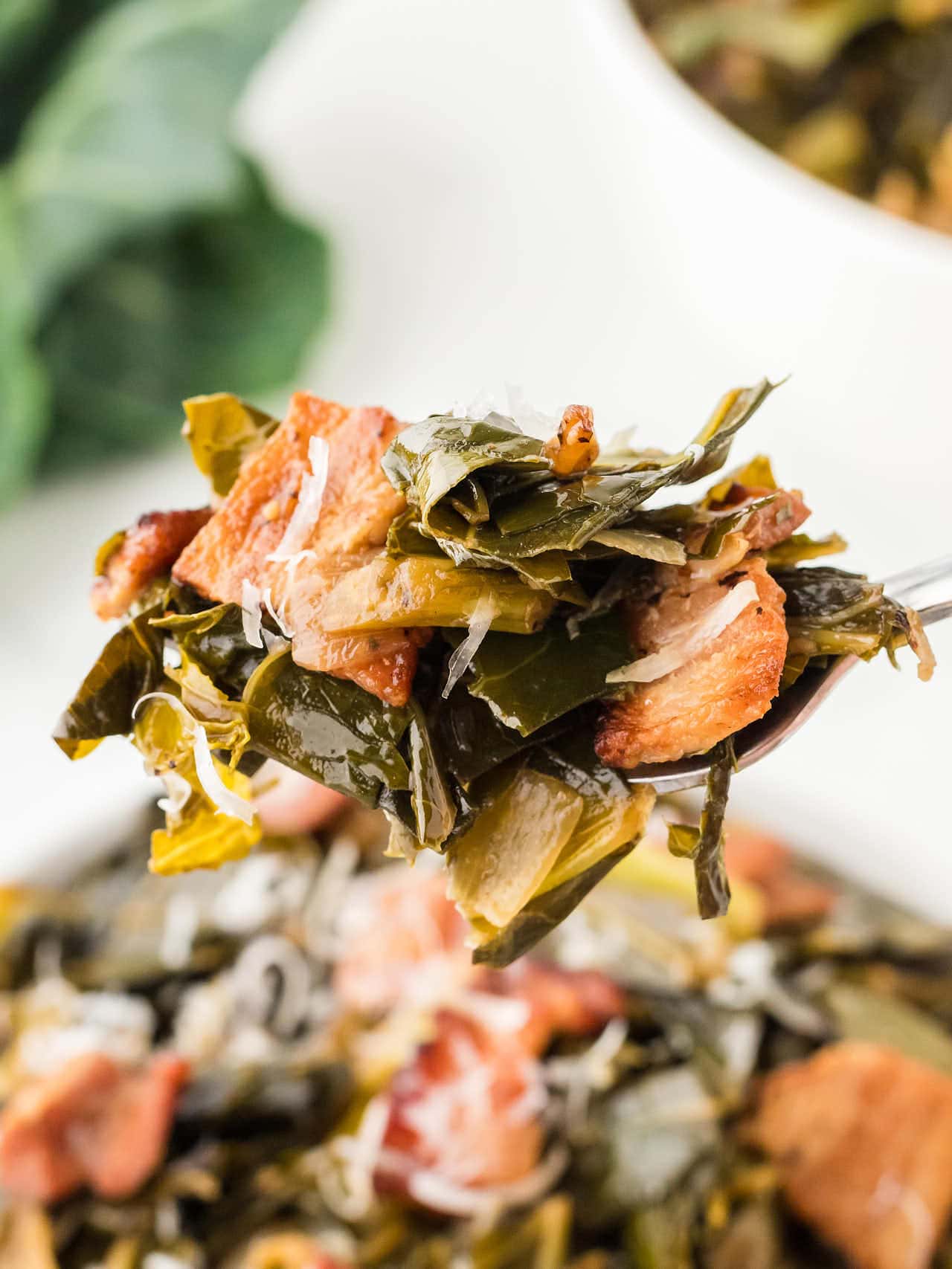 Close up of tender collard greens with crispy bacon and onions lifted on a spoon, showing savory texture and flavor.