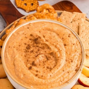Pumpkin dip in a white bowl topped with cinnamon, surrounded by vanilla wafers, graham crackers, and pretzels.
