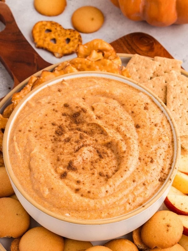 Pumpkin dip in a white bowl topped with cinnamon, surrounded by vanilla wafers, graham crackers, and pretzels.