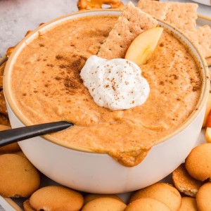 Pumpkin dip with whipped cream, apple slice, and graham cracker garnish in a white bowl with cookies and pretzels.