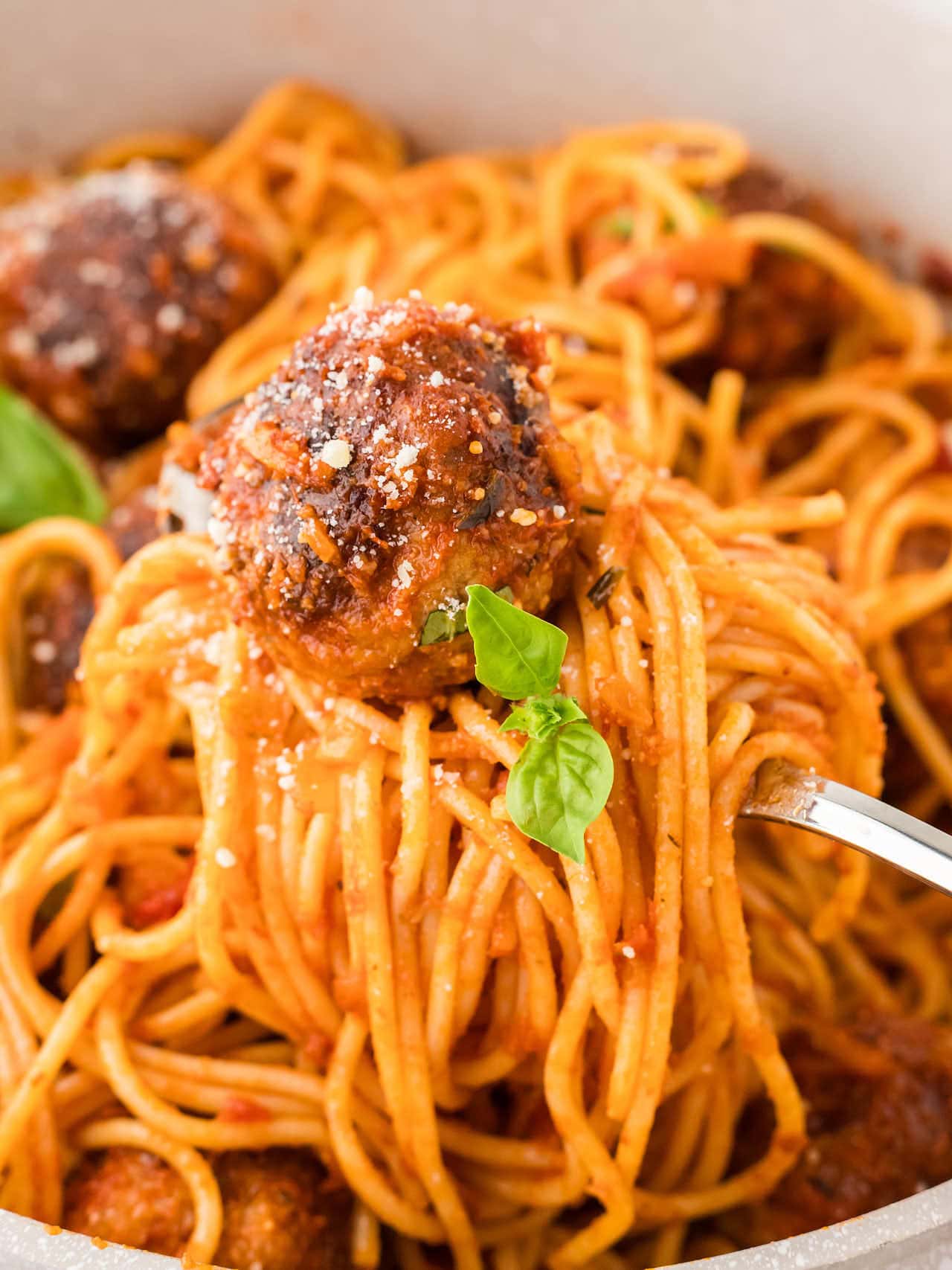 Close-up of Spaghetti and Meatballs twirled on a fork with marinara sauce, Parmesan cheese, and a fresh basil garnish.