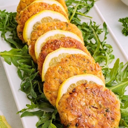 Crispy golden salmon patties served on fresh arugula with lemon slices on a white platter.