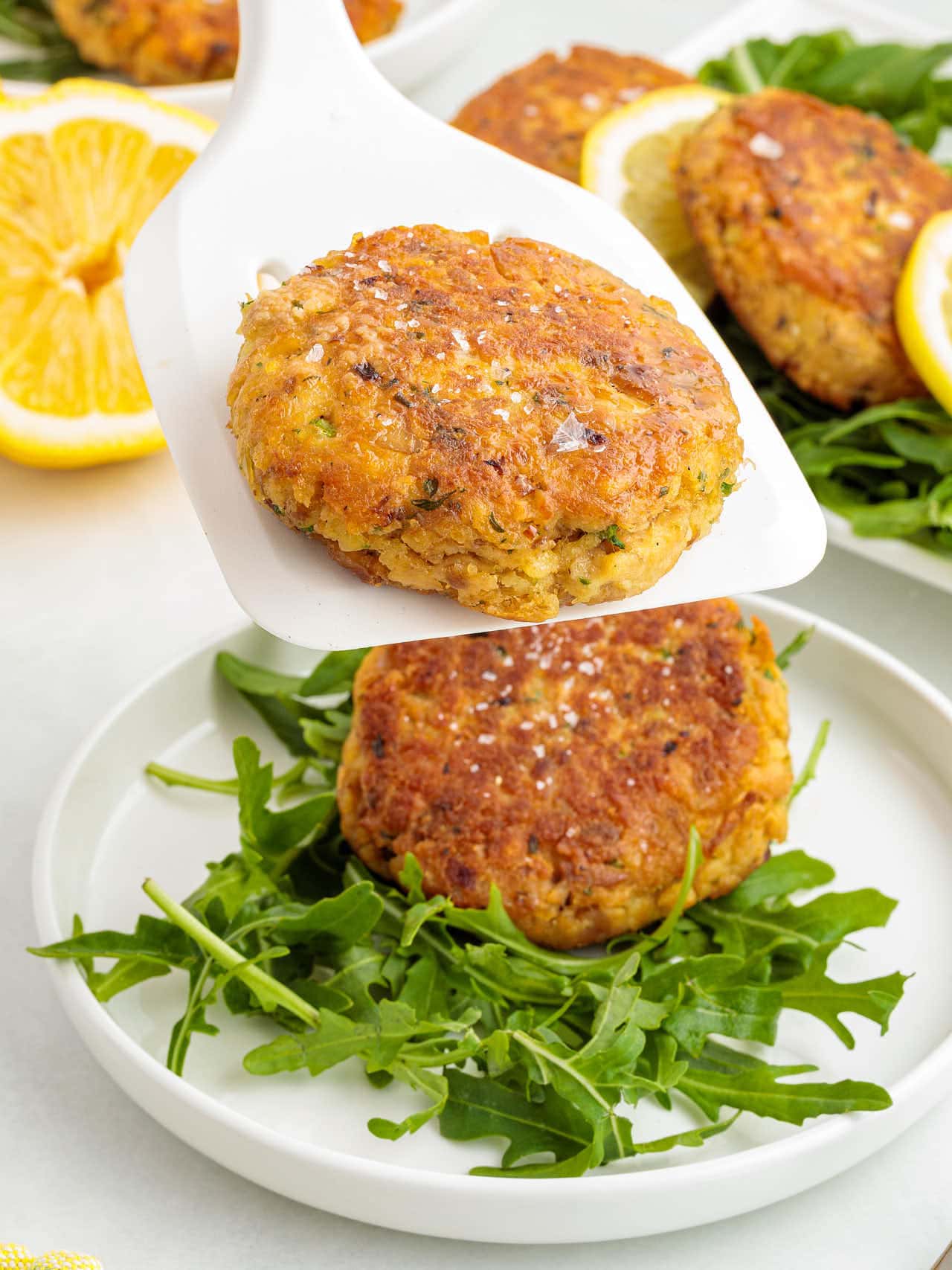 Crispy salmon patty being lifted with a white spatula above a plate of arugula.
