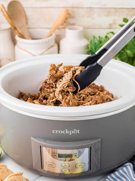 Slow cooker carnitas with crispy edges lifted from a crock pot with tongs.