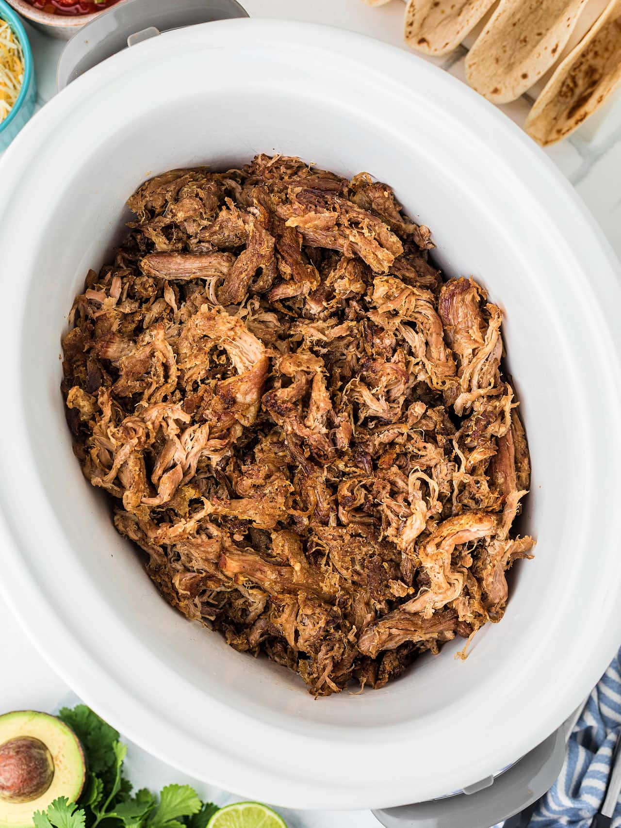 Slow cooker carnitas surrounded by tortillas, salsa, and avocado.