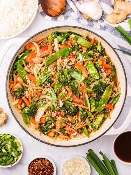 Overhead view of Ground Chicken Stir Fry in a white skillet next to a bowl of steamed rice, garlic, and fresh ginger.