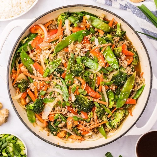 Overhead view of Ground Chicken Stir Fry in a white skillet next to a bowl of steamed rice, garlic, and fresh ginger.