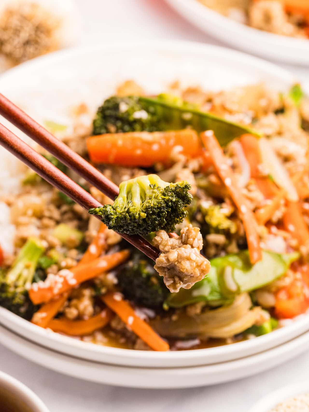 Chopsticks holding broccoli and ground chicken above a bowl of Ground Chicken Stir Fry served over rice.