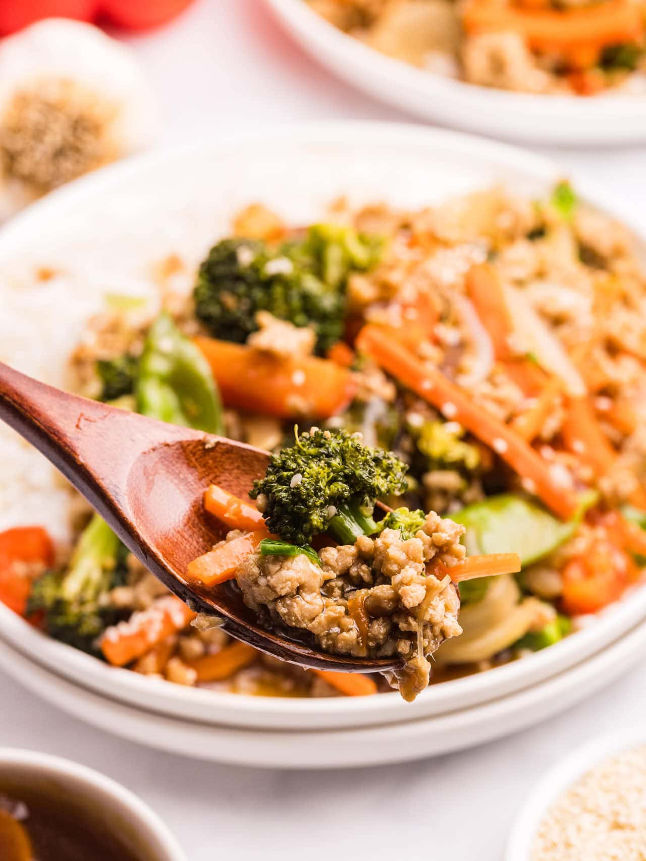 A wooden spoon lifting a scoop of Ground Chicken Stir Fry with broccoli, carrots, and ground chicken over a plate.