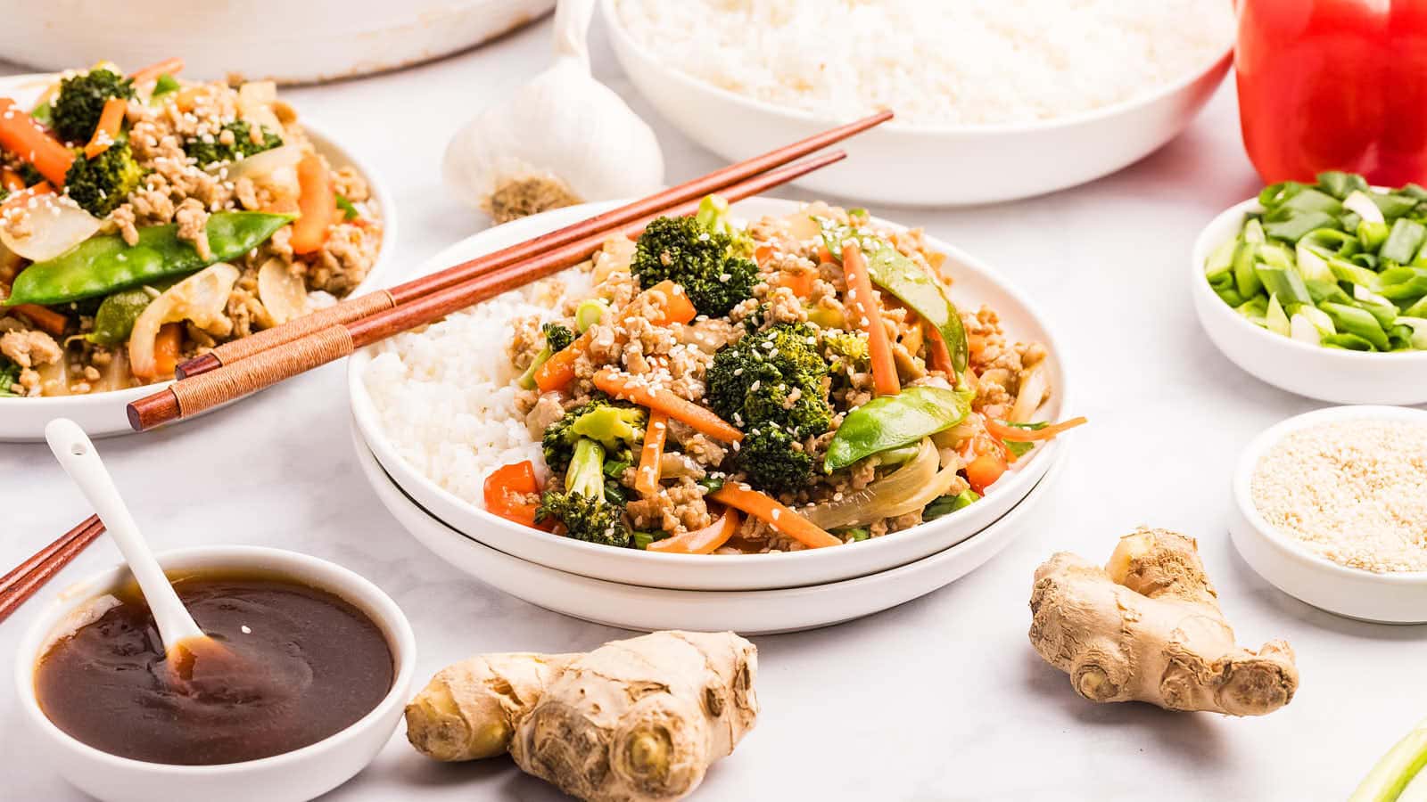 Two plates of Ground Chicken Stir Fry served over white rice with a small bowl of sauce and fresh ginger nearby.