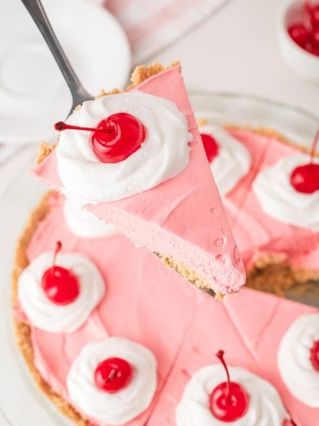Slice of kool aid pie being lifted from the whole pie with a server showing the pink filling and graham crust.