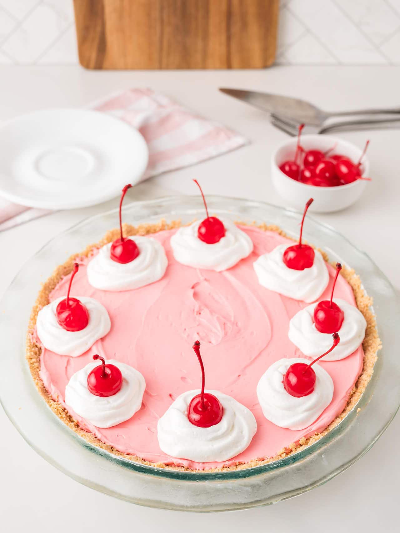 Whole kool aid pie with cool whip rosettes and maraschino cherries with serving plates nearby.