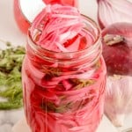 Tall mason jar of homemade pickled red onions with a fork lifting a portion out, fresh herbs and red onion nearby.