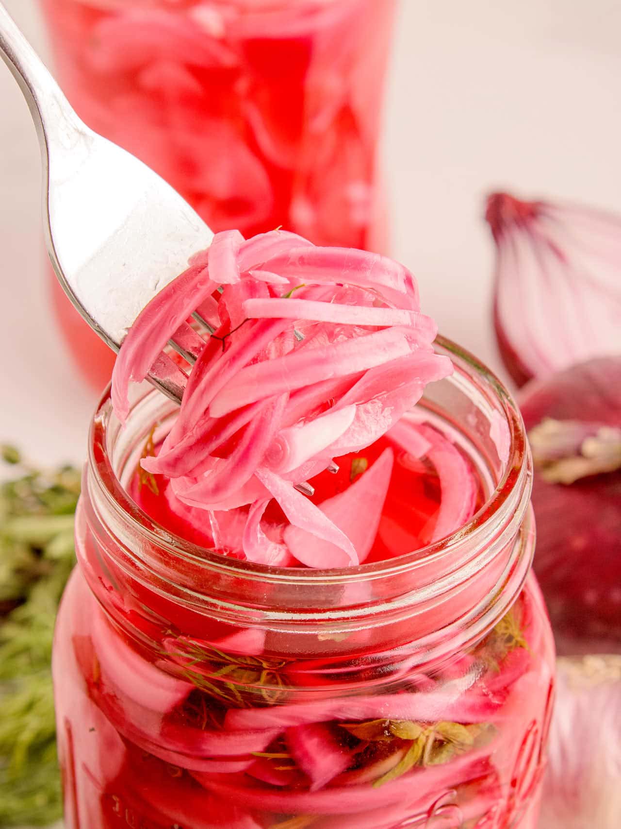 Fork pulling a tangle of pink pickled red onion ribbons from a mason jar with raw red onion in the background.
