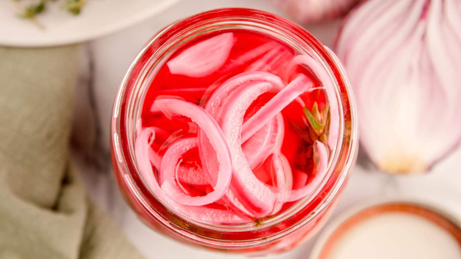 Overhead view of pickled red onions in a mason jar with bright pink brine, sliced onions, and fresh herb sprigs.