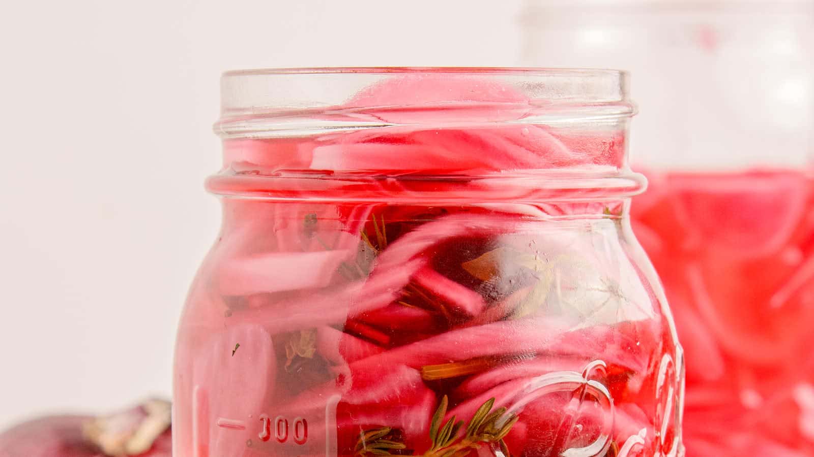 Side view of a mason jar of pickled red onions with a fresh thyme sprig floating in bright pink vinegar brine.