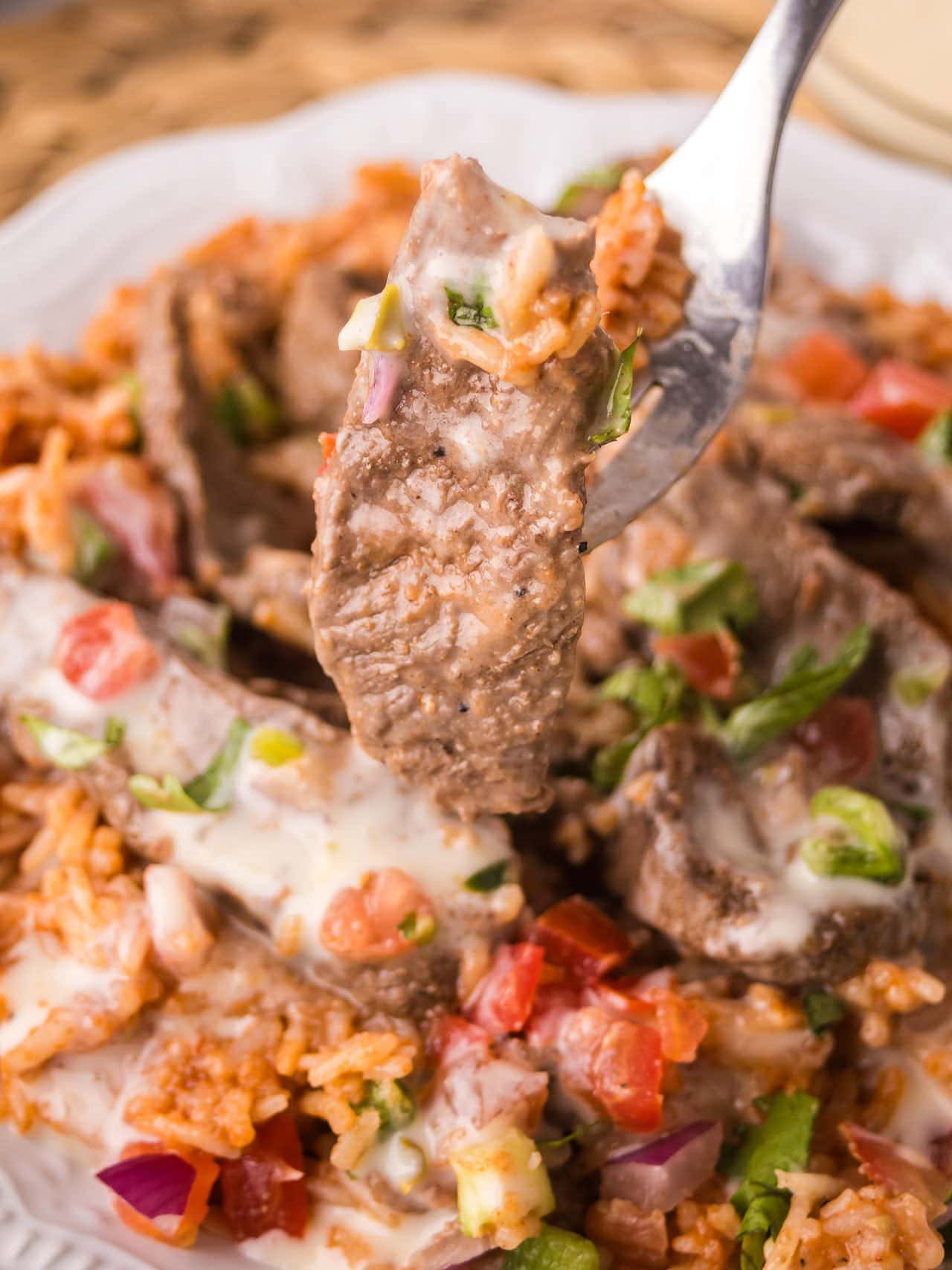 Fork lifting a tender bite of Steak and Rice with white queso and pico de gallo from a serving plate.