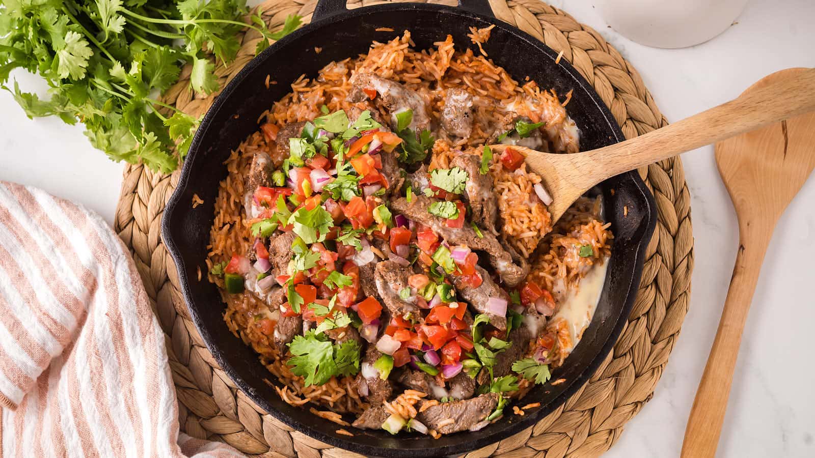Steak and Rice in a cast iron skillet with a wooden spatula, topped with pico de gallo, white queso, and cilantro.