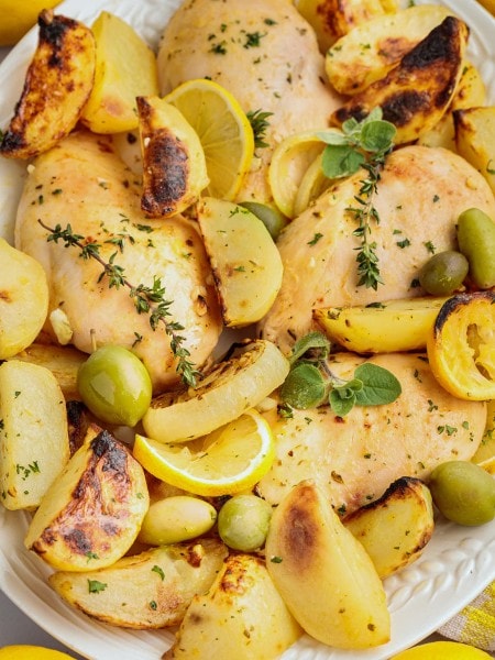 Greek Lemon Chicken and Potatoes on a white platter with lemon slices, green olives, and fresh herbs.