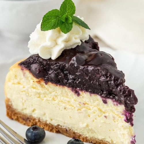 A slice of Blueberry Cheesecake topped with whipped cream and a mint sprig, with a fork and fresh blueberries.